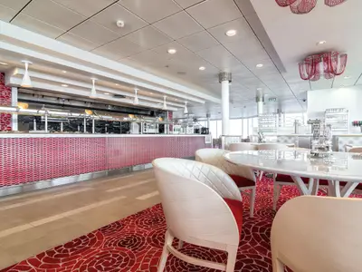 Ein helles und modernes Buffet-Restaurant an Bord der Mein Schiff 4. Im Vordergrund sind Tische und Stühle mit weiß gepolsterten Rückenlehnen und roten Sitzen zu sehen. Der Boden ist mit einem roten Teppich mit floralen Mustern ausgelegt. Im Hintergrund befindet sich ein Buffet mit warmen Speisen und einer rot-weiß gemusterten Verkleidung. Die Decke ist weiß mit Einbauleuchten und es gibt eine auffällige rote Deckenleuchte.