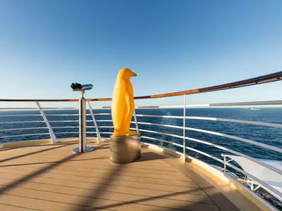 Ein gelber Pinguin-Statue und ein Fernglas stehen auf dem Teakholz-Deck der Mein Schiff 4. Im Hintergrund erstreckt sich das weite blaue Meer unter einem wolkenlosen Himmel.