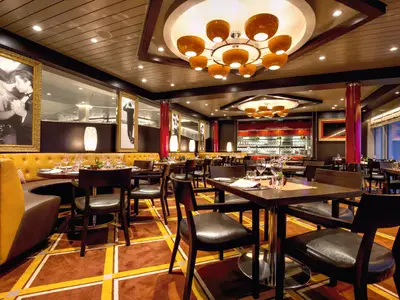 Innenansicht eines stilvollen Restaurants an Bord der Mein Schiff 4. Man sieht runde Tische mit Stühlen, gelbe Polsterbänke und große gerahmte Schwarz-Weiß-Bilder an den Wänden. Warme Beleuchtung durch Deckenleuchten und Wandlampen. Im Hintergrund ist eine Bar sichtbar. Große Fenster auf der rechten Seite geben den Blick nach draußen frei.