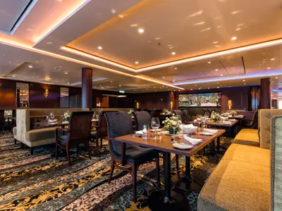 Blick in ein fein gedecktes Restaurant der Mein Schiff 4 mit Reihen von Tischen, Stühlen und bequemen Bänken. Im Hintergrund sind Wandlampen und eine große Bildtafel zu sehen. Die Decke ist mit indirekter Beleuchtung und Spots ausgestattet.