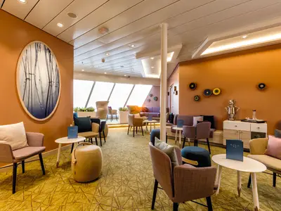 Eine moderne Lounge an Bord der Mein Schiff 4 mit gemütlichen Sesseln und Sofas in Lila- und Beigetönen, dazu kleine weiße Tische. Die Wände sind orange und hellbraun gestrichen und mit ovalen Bildern und runden Dekorationen versehen. Große Fenster auf der linken Seite bieten Ausblick nach draußen. Ein silberner Samowar steht auf einem Sideboard.