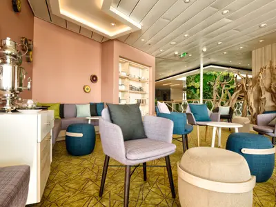 Eine moderne und gemütliche Lounge an Bord der Mein Schiff 4. Im Vordergrund stehen mehrere Sessel und Hocker in verschiedenen Blau- und Lilatönen auf einem gelb-grün gemusterten Teppich. Im Hintergrund sind eine rosafarbene Wand mit Wanddekorationen und eine große Fensterfront zu sehen, hinter der eine begrünte Wand und Holzskulpturen die Natur ins Innere holen. Ein großer, silberner Samowar steht auf einer weißen Theke links im Bild.