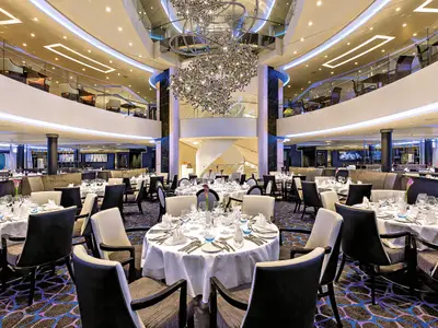 Ein Panoramafoto des stilvollen Restaurants Atlantic an Bord der Mein Schiff 4. Weiße Tische sind elegant gedeckt, umgeben von dunklen Stühlen. Eine Galerie-Ebene mit weiteren Sitzgelegenheiten ist sichtbar, und in der Mitte hängt ein großer, funkelnder Kronleuchter über einem Treppenaufgang. Die gesamte Szene ist hell und einladend beleuchtet.