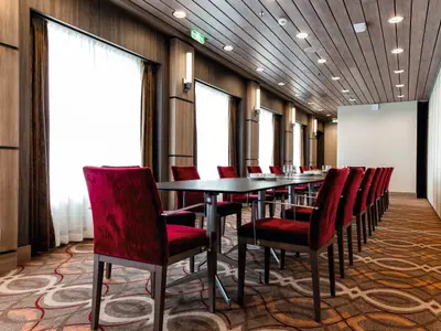 Blick in einen eleganten Konferenzraum an Bord der Mein Schiff 4. Lange Tische sind mit Wassergläsern und Tassen gedeckt. Rote Samtstühle stehen bereit. Die Wände sind holzvertäfelt, die Decke mit Holzlatten verkleidet. Helle Fenster mit braunen Vorhängen und LED-Beleuchtung sorgen für eine angenehme Atmosphäre.