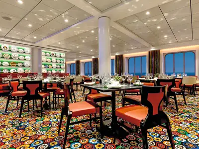 Ein elegantes, helles Restaurant an Bord der Mein Schiff 4 mit zahlreichen gedeckten Tischen. Grosse Fenster bieten einen weiten Blick auf das offene Meer. Die Wände sind mit Regalen voller dekorativer Teller geschmückt. Der Teppichboden ist auffällig gemustert in bunten Farben wie Orange, Gelb, Grün und Blau.