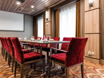 Blick in einen Konferenzraum an Bord der Mein Schiff 4. Eine lange Holztafel mit vielen roten Stühlen ist mittig platziert. Auf der linken Seite befindet sich eine große weiße Projektionsfläche an der Wand. Rechts sind hohe Fenster mit hellen Vorhängen und Holzvertäfelung zu sehen.