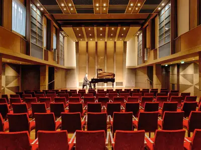 Ein Pianist im Anzug spielt auf einem Flügel auf einer Holzscheune. Die Bühne ist beleuchtet, und im Vordergrund sind Reihen von leeren, samtigen roten Stühlen zu sehen. Die Wände des Saals sind aus Holz mit vertikalen Paneelen und Fenstern. Eine elegante Konzertatmosphäre an Bord der Mein Schiff 4.