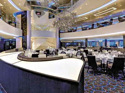 Ein Panoramaaufnahme des Hauptrestaurants an Bord der Mein Schiff 4. Im Vordergrund sind elegant gedeckte runde Tische mit weißen Tischdecken und schwarzen Stühlen zu sehen. Im Hintergrund erstreckt sich das Restaurant über zwei Etagen, die durch eine geschwungene Treppe und Geländer verbunden sind. Eine imposante, funkelnde Kronleuchter hängt von der Decke. Große Fenster im hinteren Bereich bieten einen Blick auf das Meer.