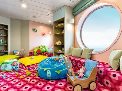 Ein bunter Spielbereich an Bord der Mein Schiff 4 mit pinkem Teppich, Sitzsäcken und Holzregalen voller Spielzeug. Ein großes Bullauge bietet Ausblick auf das Meer und entfernte Berge.