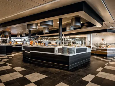 Blick in ein modernes Buffet-Restaurant an Bord der Mein Schiff 4. Mehrere Theken mit Speisen unter silbernen Lampen sind zu sehen. Im Hintergrund steht ein Koch in weißer Uniform vor einer gefliesten Wand.