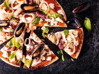 A close-up of a partially sliced seafood pizza with mussels, calamari rings, octopus pieces, shrimp, and chunks of white cheese, garnished with basil leaves, on a dark, textured surface, possibly an Bord der Mein Schiff 4. Additional mussels and basil leaves are scattered around the pizza.