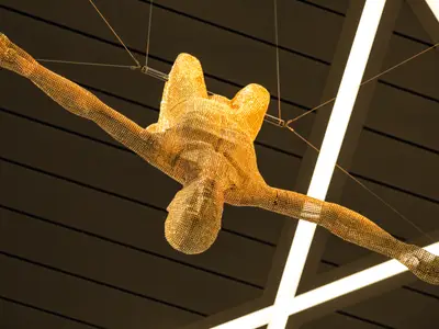 Blick von unten auf eine goldene Skulptur eines Menschen mit ausgestreckten Armen und Beinen, als würde er springen oder schweben. Sie ist aus unzähligen kleinen, goldenen Elementen geformt und hängt von der Decke der Mein Schiff 4. Im Hintergrund sind dunkle Deckenplatten und helle, leuchtende Streifen zu sehen.