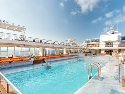 Blick auf das obere Deck der Mein Schiff 4 mit zwei langen, miteinander verbundenen Pools. Orangefarbene Liegeflächen säumen die Pools. Im Hintergrund ist das blaue Meer zu sehen, darüber ein heller Himmel. Auf der oberen Ebene des Decks gibt es weitere Liegestühle.