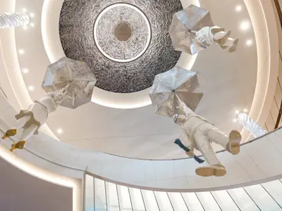 Blick nach oben im Atrium der Mein Schiff 4, wo vier schwebende, menschliche Figuren mit silbernen Regenschirmen von der Decke hängen. Die Decke hat eine zentrale, kreisförmige Struktur mit einem Reliefmuster und umlaufender Beleuchtung. Die Figuren sind in hellen Farben gehalten, zwei tragen goldene Schuhe, einer eine dunkle Brille und einen blauen Gegenstand in der Hand. Die Szene ist hell und modern.