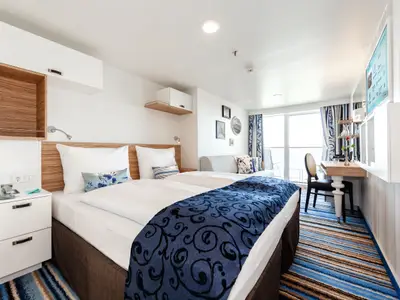 Ein geräumiges und modern eingerichtetes Hotelzimmer mit zwei Einzelbetten, die zu einem Doppelbett zusammengeschoben sind, an Bord der Mein Schiff 4. Ein dunkelblaues, gemustertes Plaid liegt über dem Fußende der Betten. Links vom Bett steht ein Nachttisch mit Telefon und Steckdosen. Rechts im Bild sind ein Schreibtisch mit Stuhl und ein Wandregal mit Fernseher zu sehen. Große Glas-Schiebetüren führen zu einem Balkon. Der Boden ist mit einem blau-braun gestreiften Teppich ausgelegt.