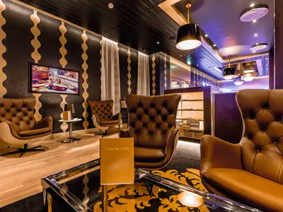 Eine luxuriöse Lounge an Bord der Mein Schiff 4 mit bequemen, braunen Ledersesseln. Die Wände sind in Schwarz mit wellenförmigen goldenen Streifen gehalten. Ein großer Fernseher zeigt eine Aufnahme von Zigarren und einem Glas Whiskey. Der Boden ist teils aus hellem Holz, teils mit einem dunklen Teppich mit goldenen Ornamenten ausgelegt. Verschiedene Beleuchtungselemente, darunter Hängelampen, erhellen den Raum.