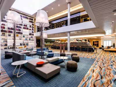 Blick in eine weitläufige Lounge auf der Mein Schiff 4, mit blauer Teppichware, modernen Sitzmöbeln und einem hohen Bücherregal. Im Hintergrund ist ein Schiffsmodell zu sehen.