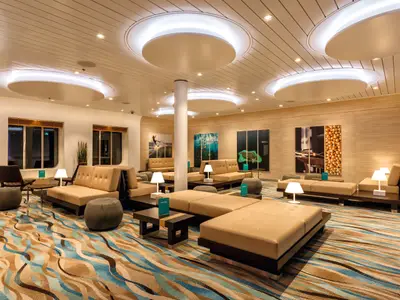 Eine moderne und gemütliche Lounge an Bord der Mein Schiff 4. Beige Polstermöbel in Reihen mit dunklen Holzgestellen stehen auf einem Teppich mit blauen und braunen Wellenmustern. Die Decke hat runde, beleuchtete Elemente, und an den Wänden hängen Kunstwerke. Große Fenster auf der linken Seite bieten Ausblick.