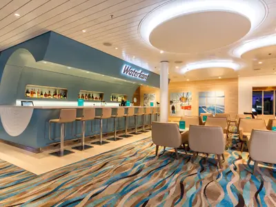 Ein geräumiger und modern gestalteter Bar- und Loungebereich an Bord der Mein Schiff 4. Eine lange, türkisblaue Bar mit Nischen voller Spirituosen und hohen Barhockern steht links. Rechts gruppieren sich helle Sessel um niedrige Tische auf einem Teppich mit Wellenmuster. An der Wand hängen Bilder mit winterlichen Meeresmotiven, und die Decke ist mit runden Lichtelementen verziert.