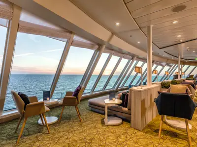 Ein Panoramafoto einer geräumigen Lounge an Bord der Mein Schiff 4. Große Fensterfronten bieten einen weiten Blick auf das offene Meer und den Horizont, wo sich die Sonne senkt. Im Vordergrund befinden sich moderne Sitzgelegenheiten wie Sessel und Sofas mit weichen Kissen, teilweise um kleine Beistelltische gruppiert. Die Beleuchtung ist gedämpft und warm. Die Einrichtung ist in hellen Holz- und Stofftönen gehalten. Der Boden ist mit einem gemusterten Teppich in Grüntönen ausgelegt.