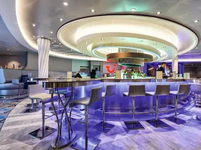 Innenansicht der TUI Bar auf der Mein Schiff 4. Eine geschwungene Bar mit blauer Beleuchtung steht im Mittelpunkt, dahinter ein Barkeeper. Elegante Hocker und Sitzbereiche sind sichtbar.