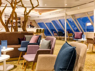 Ein Barkeeper mixt Drinks hinter einer hellen Bar in einer modernen Lounge an Bord der Mein Schiff 4. Gäste sitzen auf bunten Stühlen und Sofas mit Kissen, dahinter sind große, schräge Fenster mit Blick auf das blaue Meer.