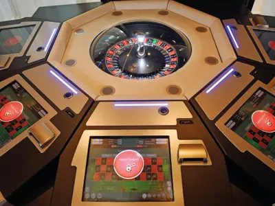 Blick von oben auf ein modernes, elektronisches Roulette-Spiel an Bord der Mein Schiff 4. Mehrere Spielerstationen mit Touchscreens sind um ein zentrales, rotierendes Roulette-Rad angeordnet. Die Bildschirme zeigen ein "Station locked!"-Symbol und das Spielfeld mit Zahlen und Wettoptionen. Blaue LED-Lichter beleuchten die Spielerbereiche.