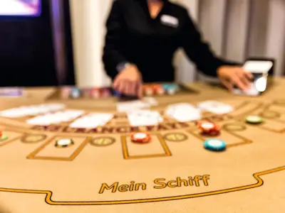 Blick auf einen Black Jack-Tisch an Bord der Mein Schiff 4. Im Vordergrund liegen Spielkarten und Pokermarker. Die Hände eines Croupiers verteilen im Hintergrund weitere Karten.