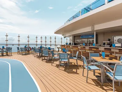 Blick auf das Außendeck der Mein Schiff 4 mit der 'Überschaubar', Tischen und blauen Stühlen. Im Vordergrund ein blauer Joggingpfad, im Hintergrund die weite See und blauer Himmel.