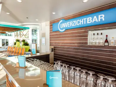 Ein breiter Blick auf eine moderne Bar an Bord der Mein Schiff 4. Im Vordergrund stehen zahlreiche umgedrehte Weinglaeser auf der Theke. Im Hintergrund ist eine Holzvertäfelung mit einem blauen Schild mit der Aufschrift „UNVERZICHTBAR“. Darunter ist eine Öffnung mit Spirituosenflaschen zu sehen. Links der Bar befinden sich orangefarbene Liegestühle am Pooldeck.