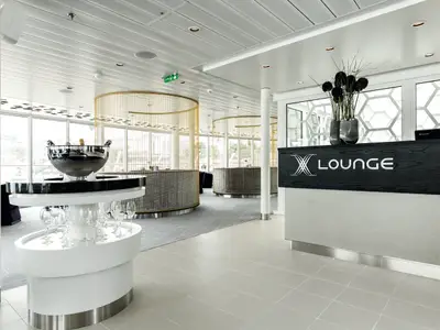 Innenansicht der X Lounge auf der Mein Schiff 4. Im Vordergrund ein Empfangstresen mit "X Lounge"-Schriftzug und Blumen. Links eine Weinkühl-Bar. Im Hintergrund zwei runde Bartresen und große Fenster mit Meerblick.