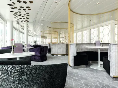 Die helle und moderne X-Lounge an Bord der Mein Schiff 4. Im Vordergrund sind Tische mit lilafarbenen Menükarten zu sehen, umgeben von schwarzen und lilafarbenen Sesseln. Eine goldene Perlenkette trennt den Sitzbereich vom dahinterliegenden Barbereich. Große Fensterfronten lassen viel Licht herein und bieten einen Blick nach draußen.