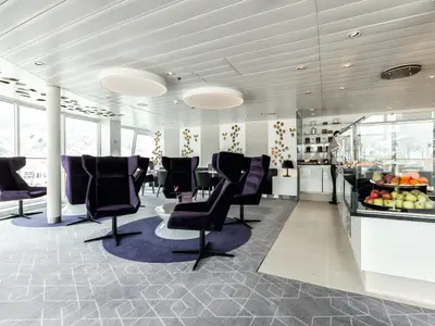 Blick in eine moderne Lounge an Bord der Mein Schiff 4 mit violetten Sesseln, großen Fenstern und einer Bar, die frisches Obst präsentiert. Die Wände sind mit dekorativen Blattformen verziert.