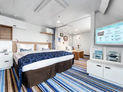 Blick in eine Kabine an Bord der Mein Schiff 4. Im Vordergrund ein Doppelbett mit blauer Tagesdecke. Rechts daneben ein Wohnbereich mit Sofa, Couchtisch und Flachbildfernseher an der Wand.