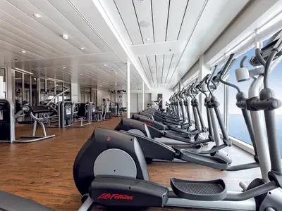 Blick in den Fitnessraum der Mein Schiff 4 mit einer Reihe von Ellipsentrainern am Fenster mit Meerblick. Im Hintergrund sind weitere Fitnessgeräte zu sehen, darunter Kraftstationen. Der Boden ist aus Holz, und die Decke ist hell und mit eingelassenen Lichtern versehen.