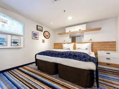Blick in eine Kabine an Bord der Mein Schiff 4. Man sieht ein großes Bett mit weißen Kissen und einer blau gemusterten Decke. Daneben steht ein kleiner weißer Nachttisch mit Holzapplikationen. Gegenüber hängt ein großer Flachbildfernseher mit "Mein Schiff"-Branding und darunter ein Regal mit Broschüren. Der Teppichboden ist gestreift in Blau-, Braun- und Weißtönen.