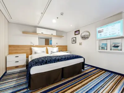 Innenansicht eines Kabinenzimmers auf der Mein Schiff 4. Ein großes Doppelbett mit blauer Tagesdecke dominiert den Raum. Auf der rechten Seite hängt ein Flachbildfernseher über einem Regal mit Broschüren. Die Wände sind weiß, der Teppich gestreift in Blau- und Brauntönen.
