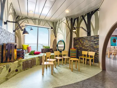 Blick in den liebevoll gestalteten Kinderbereich der Mein Schiff 4 mit Wandmalereien von Bäumen. Helle Holzstühle stehen vor einer Sitzbank mit Blattmuster und bunten Kissen. Ein großes Fenster gibt den Blick aufs Meer frei.