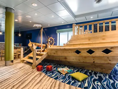Ein weitläufiger Spielbereich für Kinder an Bord der Mein Schiff 4, dominiert von einem großen, liebevoll gestalteten Holzboot mit Steuerrad und Treppen. Der Boden ist mit einem blauen Wellenteppich und einem sandfarbenen Teppich mit Strandmuster bedeckt. Links sind kleine Holzstühle und ein grüner Pfeiler sichtbar. Ein großes Fenster im Hintergrund lässt Tageslicht herein. Zwei rote Würfel und ein Buch liegen auf dem Teppich. Die Wände sind dunkelblau gestrichen.