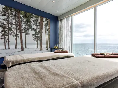 Ein luxuriöser Spa-Raum an Bord der Mein Schiff 4 mit zwei Massageliegen und Blick auf das weite Meer durch eine große Fensterfront. An der Wand befindet sich ein großes Wandbild von Kiefern in einer verschneiten Landschaft. Auf einem kleinen Beistelltisch stehen Massageöle bereit. Auf der vorderen Liege liegen braune Handtücher und ein Stück Treibholz.