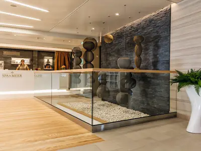Eine Frau in Arbeitskleidung steht lächelnd hinter einer beleuchteten, weißen Rezeption mit der Aufschrift „SPA & MEER“ an Bord der Mein Schiff 4. Im Hintergrund sind dunkle Steinwände und Holzpaneele zu sehen. Vor der Rezeption befindet sich eine gläserne Abtrennung mit weißen Zierkieseln.