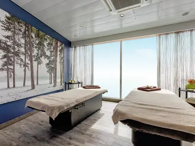 Ein luxuriöser Spa-Behandlungsraum auf der Mein Schiff 4 mit zwei Massageliegen, einem großen Wandbild mit Winterwald und bodentiefen Fenstern, die einen weiten Blick auf das Meer bieten. Warme, dezente Beleuchtung und natürliche Farbtöne schaffen eine entspannte Atmosphäre.