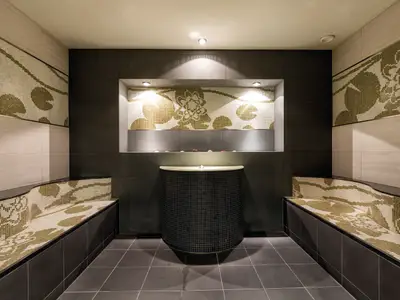 Ein luxuriöses Dampfbad an Bord der Mein Schiff 4 mit kunstvollen Mosaikwänden, die florale Muster in Gold- und Beigetönen zeigen. Die geschwungenen Sitzbänke sind ebenfalls mit passenden Mosaiken verziert. In der Mitte befindet sich eine runde Theke mit einem Becken, darüber eine Nische mit weiteren Mosaiken und indirekter Beleuchtung. Der Raum ist in dunklen und hellen Erdtönen gehalten und strahlt Ruhe und Eleganz aus.