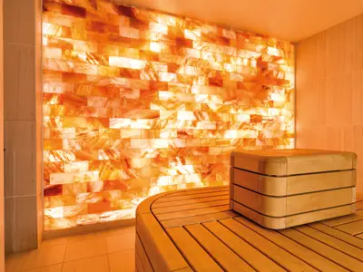Ein Innenraum an Bord der Mein Schiff 4 mit einer leuchtenden Wand aus rosafarbenen Salzziegeln. Davor steht eine geschwungene Holzbank mit einem würfelförmigen Holz-Beistelltisch.