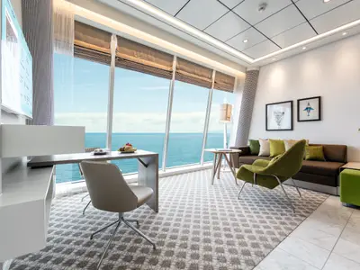 Blick in eine helle, modern eingerichtete Kabine der Mein Schiff 4 mit raumhohen Fenstern und Meerblick. Links ein Schreibtisch mit Stuhl, daneben eine Sitzecke mit dunkelbraunem Sofa und hellgrünen Kissen sowie einem olivgrünen Sessel. An der Wand hängen zwei gerahmte Bilder.
