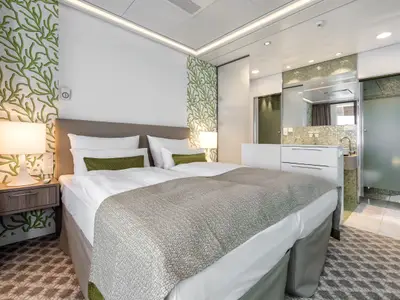 Blick in eine helle Doppelbett-Kabine der Mein Schiff 4. Das Bett ist weiß bezogen mit einer grauen Decke und grünen Kissen. Links vom Bett steht ein Nachttisch mit einer leuchtenden Lampe. Die Wände sind weiß mit grünen Blättermotiven. Rechts vom Bett ist ein offenes Badezimmer mit Doppelwaschbecken und Spiegeln.
