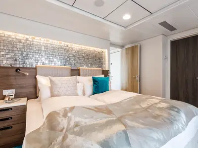 Dieses Foto zeigt ein geschmackvoll eingerichtetes Schlafzimmer an Bord der Mein Schiff 4. Im Vordergrund ein großes Doppelbett mit weißen Laken und einem aquafarbenen Kissen. Über dem Kopfteil ist eine dekorative Wand aus kleinen, spiegelnden Kacheln zu sehen, die von oben beleuchtet wird. Rechts im Bild sind zwei Türen zu erkennen, eine helle Holztür im Hintergrund und eine dunkle Holztür im Vordergrund.