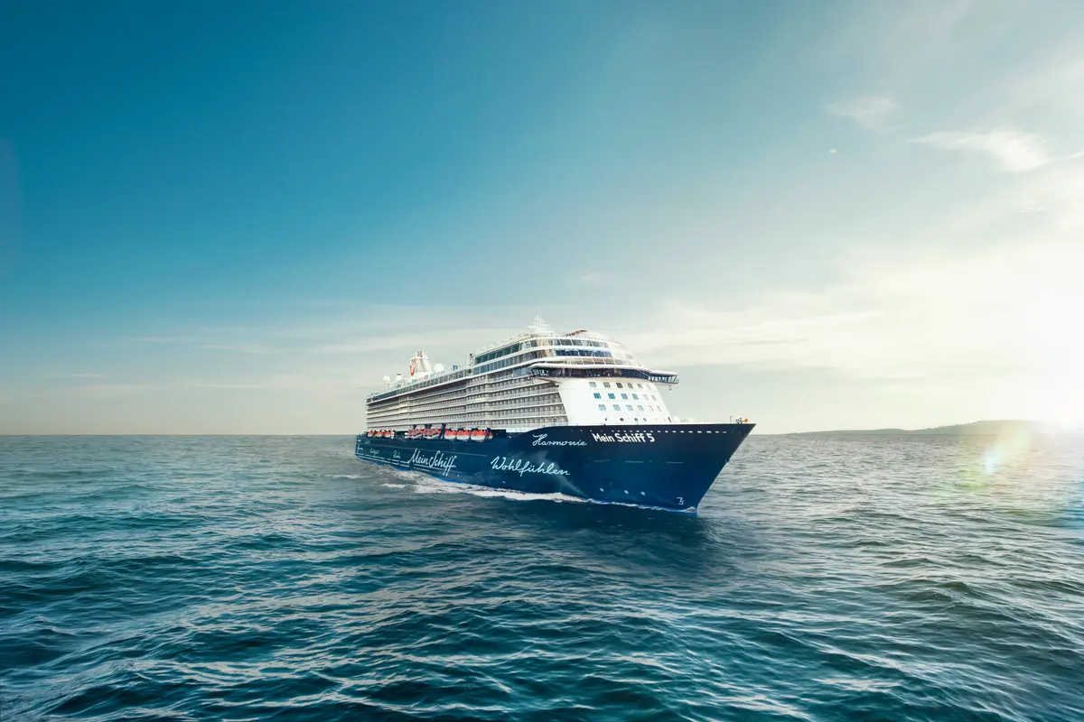 Die Mein Schiff 5 kreuzt bei strahlendem Sonnenschein auf dem tiefblauen Meer. Das moderne Kreuzfahrtschiff ist vom Bug aus schräg vorne zu sehen.