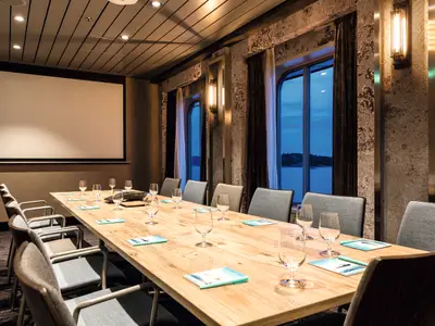 Ein gut beleuchteter Konferenzraum an Bord der Mein Schiff 5. In der Mitte steht ein langer Holztisch, gedeckt mit Weingläsern, Notizblöcken und Stiften. Graue Stühle säumen den Tisch. Links befindet sich eine große Projektionsfläche, rechts drei Fenster mit Blick auf eine bewaldete Küstenlinie und das Meer bei Dämmerung.