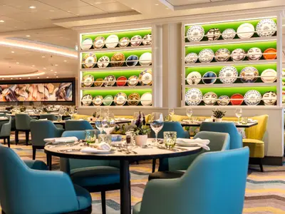 Innenansicht des Atlantik Klassik Restaurants auf der Mein Schiff 5. Zahlreiche Tische sind mit blauen und gelben Stühlen besetzt. An den Wänden hängen in Regalen Teller mit Gesichtern. Im Hintergrund ist ein großes abstraktes Gemälde zu sehen.